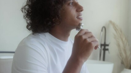 Tilt down view of biracial young man in white T-shirt massaging face with beauty roller in bathroom - Powered by Adobe