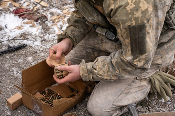 Ukrainian soldier in full camouflage uniform kneeling on the ground while loading ammunition into a...