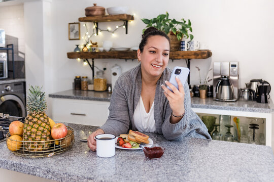 young latin woman plus size using mobile phone in kitchen at home in Mexico Latin America, hispanic overweight female 	