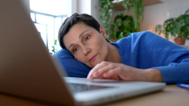 Exhausted, distracted mature woman leaning on hand while working at laptop all day. Digital and social media addiction, burnout and depression in isolation. Telecommuting, difficult remote work