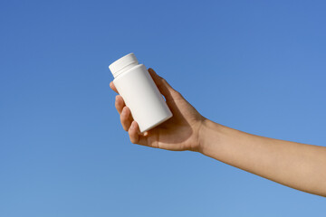 Female hand holding blank mockup pill bottle on blue sky background. Concept of pharmacy, medicine, healthcare and natural vitamins D3, magnesium, calcium, omega