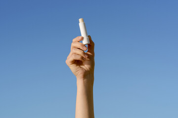 Female hand holding white lip balm mockup on blue sky background. Concept of moisturizing and...