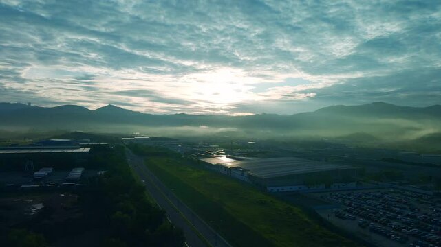 4K aerial view of an industrial area at sunrise with dramatic clouds, misty hills, and golden light, blending natural beauty and urban elements.