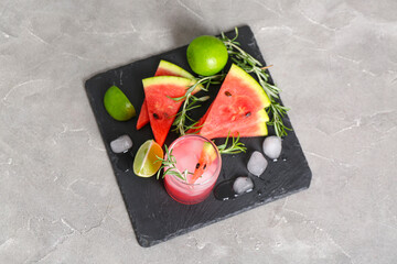 Board with glass of tasty watermelon cocktail, lime and ice cubes on grunge grey background