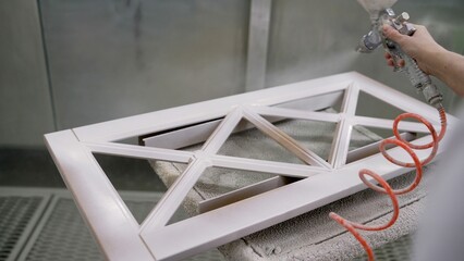 Skilled factory worker spray painting white coating onto wooden window frame using professional spray gun in industrial manufacturing setting