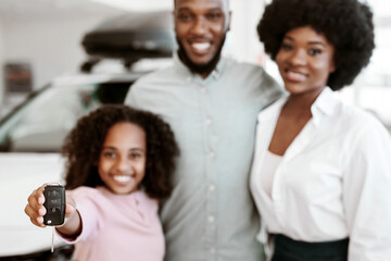 Cute black girl showing car key, standing with her parents at auto dealership, selective focus. Happy African American family purchasing new vehicle at automobile showroom, copy space