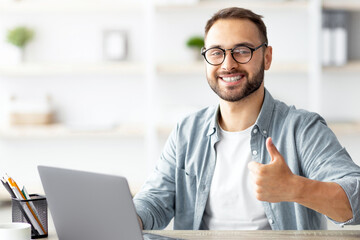 Obraz premium Happy young guy sitting at desk with flag of Turkey, using laptop, showing thumb up gesture at home. Online foreign education, emigration and citizenship, working abroad concept