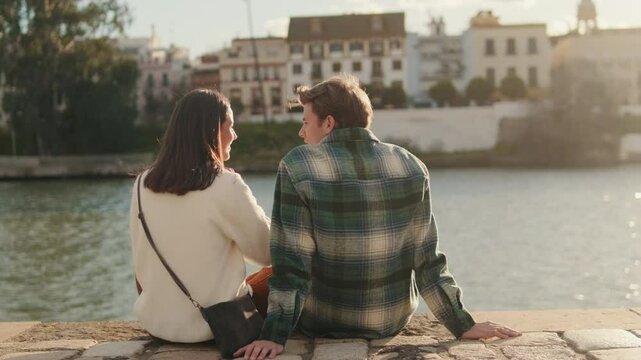 A young couple sits by the Canal de Alfonso XIII in Seville, Spain, enjoying the golden sunlight and each other's company on a warm day.