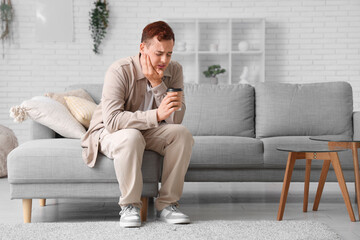 Young man with cup of coffee suffering from toothache on sofa in room