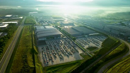 4K aerial view of an industrial facility at sunrise with factory buildings, parking lots, greenery, and misty hills, offering a serene, productive atmosphere.