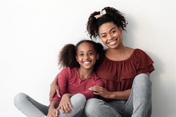 Loving black family cheerful beautiful mother and teen daughter posing on floor by empty white wall at home, wearing stylish casual outfits, embracing and smiling at camera, copy space