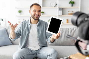 Happy male influencer filming video review of modern digital tablet on dslr camera at home, mockup on screen. Cheerful Caucasian blogger advertising modern tech product, streaming live on web