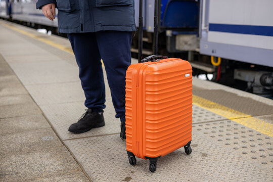 A young man with a small orange suitcase waits for a train at the train station for traveling. Lifestyle, people, adventure and tourism concept.