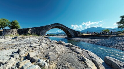 Fototapeta premium A stone bridge arches over a river with mountains in the background under a clear blue sky.