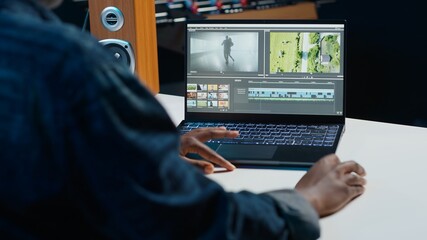 Video editor trimming segments of drone shot footage, working on promotional clip in home office. Videographer splicing film pieces, editing them using professional software on laptop, camera A