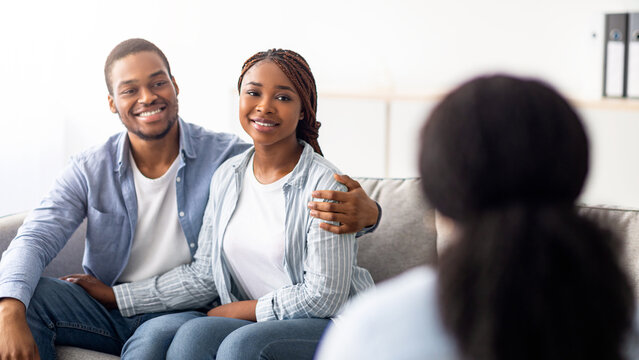 Happy married couple after successful psychotherapy at marital counselor's office. Positive young black spouses sharing results with their psychologist, hugging on couch at clinic - Powered by Adobe