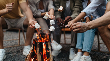 Cropped view of young people frying marshmallows on campfire, preparing yummy camping dessert outdoors, closeup. Unrecognizable youth enjoying fun summer vacation in countryside