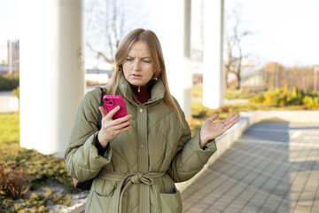 Perplexed woman holding cellphone in hands on the street, looking at screen with irritated...