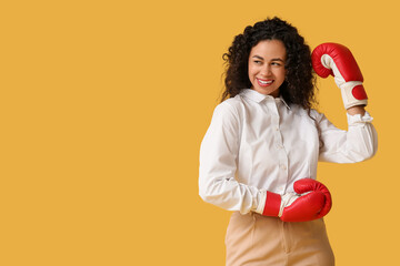 Young African-American businesswoman with boxing gloves on yellow background. Knockout concept