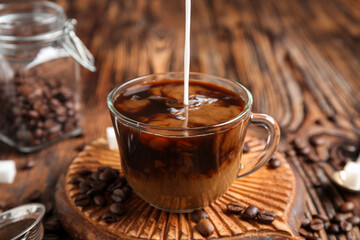 Pouring fresh milk into glass cup of hot coffee on wooden background