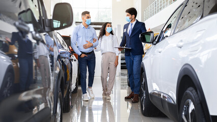 Millennial arab couple in protective face masks visiting auto showroom or exhibition while pandemic, muslim family embracing while having conversation with salesman, looking at nice white car