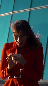 Slow motion. Euphoric sexy woman in red suit and spectacles watching her smartphone read message, rejoice winner with glossy modern business centre buiding at the background. Middle shot. Vertical