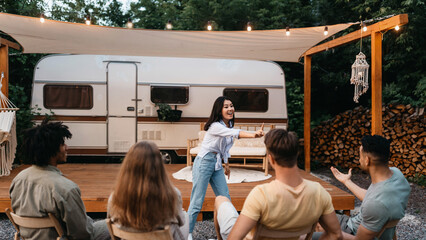 Fototapeta premium Happy Asian lady playing word guessing game with diverse friends near motorhome at campsite. Group of young people enjoying charades or pantomime riddles. Fun outdoor entertainments