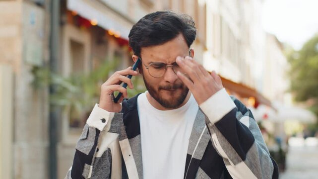Angry mad stressed Indian young man in casuals arguing while talking on smartphone irritated shout quarrel outdoors. Hispanic offended dissatisfied guy walking on urban city street. Town lifestyles.