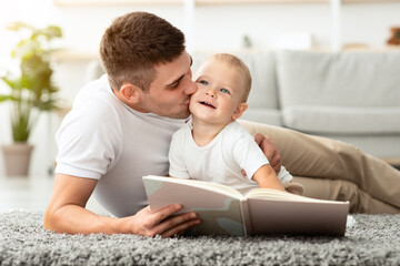 Fototapeta premium Child Care. Loving Young Father Reading Book And Kissing His Cute Toddler Son While Relaxing At Home Together, Millennial Dad Spending Time With Infant Baby, Enjoying Paternity Leave, Closeup