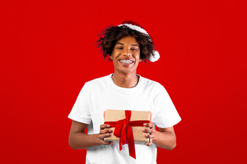 Christmas suprise. Joyful black guy in santa hat holding gift box in hands posing over blue background and smiling at camera, copy space. Positive teen enjoying presents and winter holidays