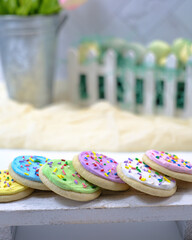 Background for personalized Easter card. Spring sugar cookies with frosting and sprinkles on wood table. Blurry backdrop with tin pail of pink tulips, white picket fence container of decorated eggs