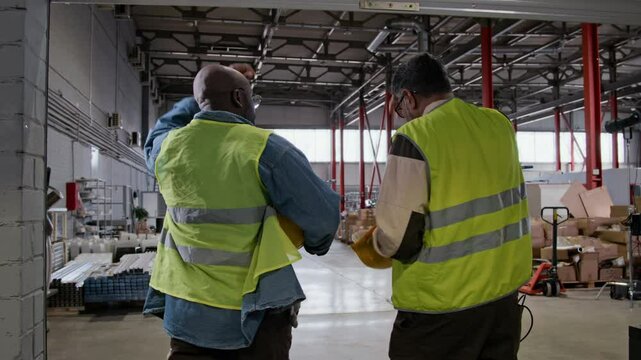 Medium full follow shot of two multiethnic male warehouse employees in uniforms, yellow safety vests arriving at work, walking in roller door, entering hangar while putting on hardhats and chatting