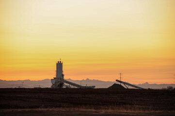 An industrial facility with conveyor systems and storage silos operates under a vibrant sunrise highlighting infrastructure development in a desert landscape