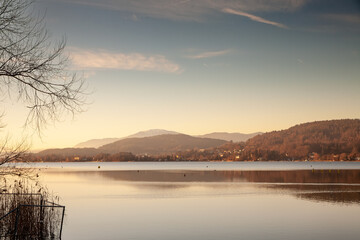 A serene evening view across the Wörthersee reveals calm waters and soft, golden hues, with distant hills framing the tranquil lakeside panorama at dusk.
