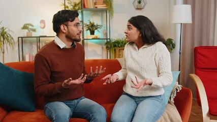 Upset young Indian couple in casual clothes arguing fighting while sitting on sofa in living room at home. Diverse disappointed girlfriend and boyfriend shouting at each other on couch in apartment.