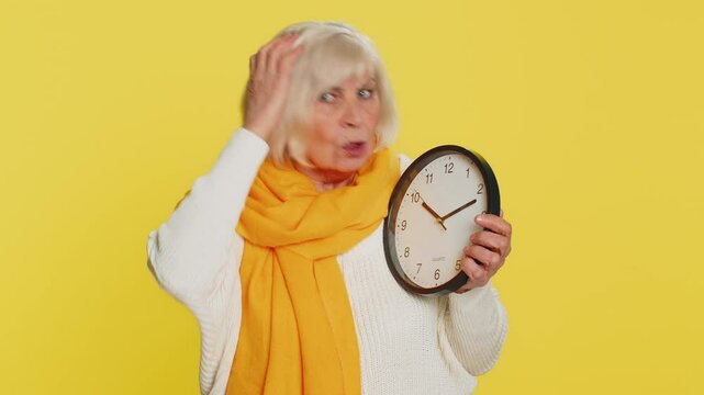 Senior old woman with anxiety checking time on clock, running late to work being in delay, deadline. Elderly grandmother looking at hour, minutes, worrying to be punctual isolated on yellow background