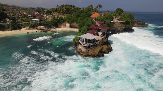 4K drone video panning around huge waves crashing against the cliffs on Nusa Ceningan near Bali in Indonesia. There is a cafe at the top of the cliffs where people can watch the large waves from.