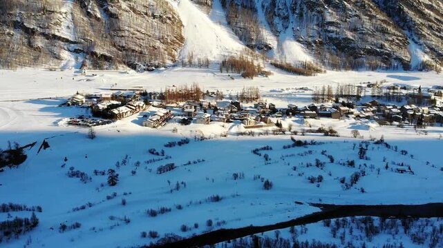 A drone pans upward, revealing a quaint village nestled at the foot of a towering mountain. The snow-covered landscape creates a serene winter scene, showcasing the harmony of the nature