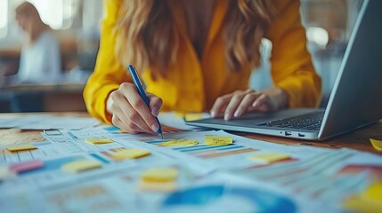 Obraz premium Close-up of a woman in a yellow shirt working with charts, sticky notes, and a laptop, symbolizing productivity, organization, and focus in a modern office environment.