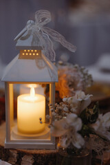 A white lantern with a candle inside is lit up. The lantern is placed on a wooden table with flowers and vases. The scene has a warm and cozy atmosphere