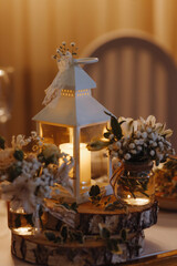A white lantern with a candle inside is lit up. The lantern is placed on a wooden table with flowers and vases. The scene has a warm and cozy atmosphere