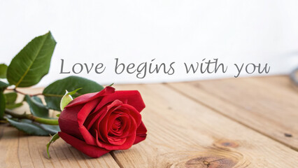 A single red rose lies on a wooden surface, with the text "Love Begins With You" displayed above. The image evokes feelings of love, romance, and hope for a new beginning