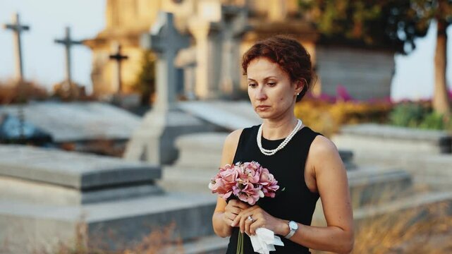 Sorrowful young woman in black dress with bouquet of flowers and handkerchief in hands, stands in cemetery. Visiting grave of deceased relative, mourning dead loved one. Appeal to God, prayer for dead