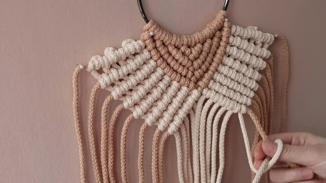 Hands carefully crafting a macrame wall hanging with beige and brown cords. Bohemian-inspired decor coming to life, highlighting the art of handmade design and the creative DIY process.
