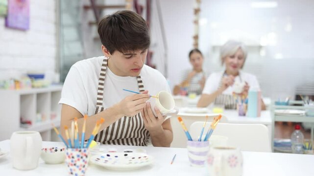 Focused young guy attending pottery class, enthusiastically painting on ceramic cup, creating unique custom design. Creative hobby concept