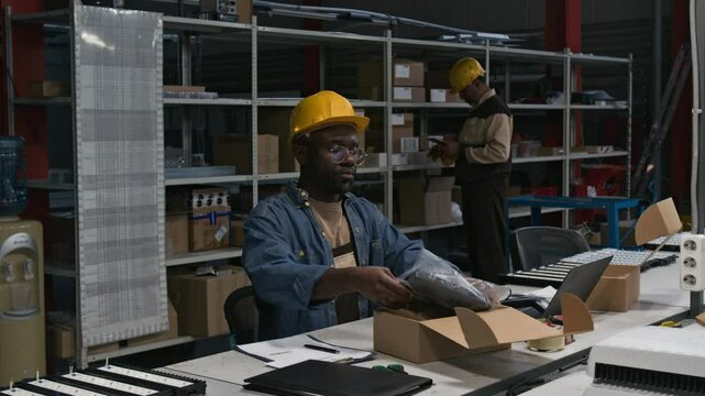 Medium shot of African American male industrial lighting factory manager in hardhat working on laptop, putting LED lamp sample in plastic bag, packing in box, colleague using tablet by product racks