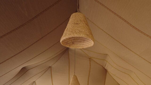 Beige canvas tent ceiling with hanging woven wicker lamps. Light illuminates the inside of the tent. Traditional Moroccan or Berber style tent interior with soft, warm lighting.