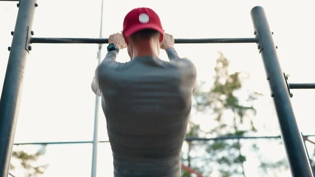 A young athletic man wearing a gray shirt and a red baseball cap performs pull-ups on a horizontal bar in a park during the day. Shot from the back. Fitness and outdoor workout concept.