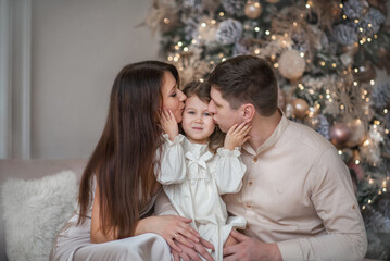 Beautiful young family - dad, mom, daughter hugging in Christmas interiors. The baby looks out the window. The girl touched her parents with her palms. The family is sitting by the window.