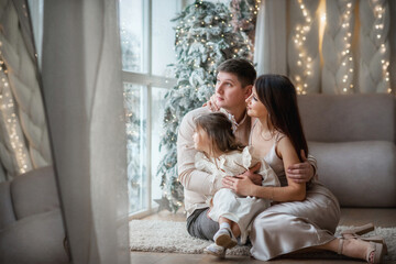 Beautiful young family - dad, mom, daughter hugging in Christmas interiors. The baby looks out the window. The girl touched her parents with her palms. The family is sitting by the window.